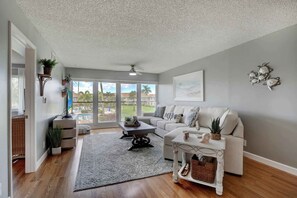 Sala de estar con sofá blanco, una mesa de centro y ventilador de techo. Se ve un ventanal con vistas a una piscina y palmeras.