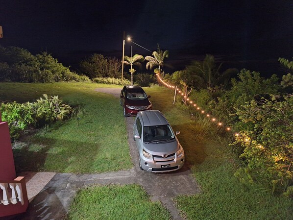 Un camino bordeado por coches aparcados e iluminado con luces colgantes, que atraviesa una zona ajardinada con palmeras y arbustos.