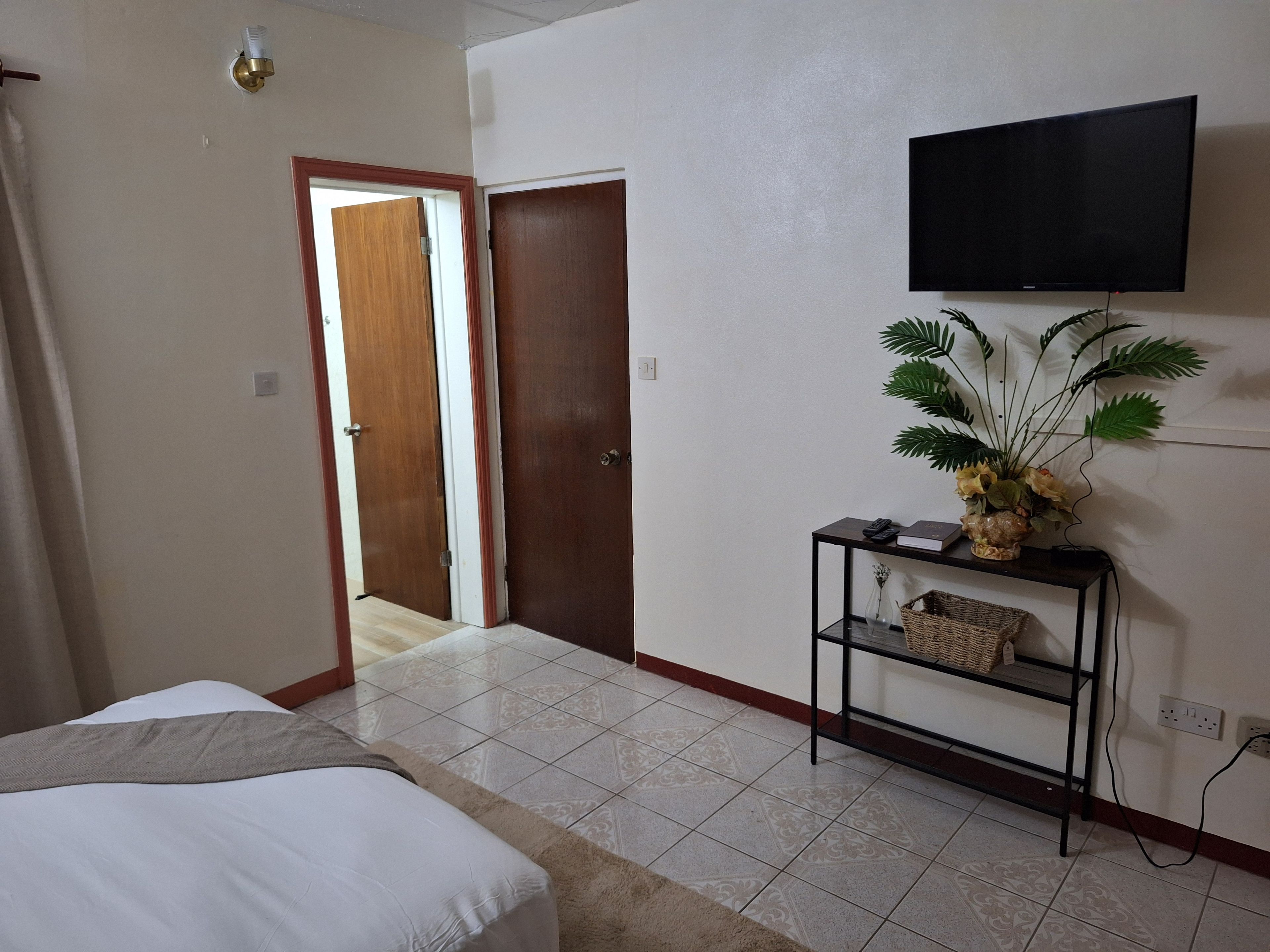 A hotel room with a bed, a television mounted on the wall, a small table with a plant, and a door leading to another room.
