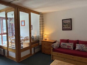 A room with a wooden sliding door leading to a dining area, a red sofa with floral cushions, a small wooden nightstand with a lamp, and a framed picture on the wall.