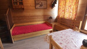 A wooden cabin room with a bed, a table, a cabinet, and a wall-mounted lamp.