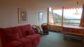 Een woonkamer met een rode bank, twee rieten fauteuils, een houten bankje en een groot raam met uitzicht op een houten terras en bergen.