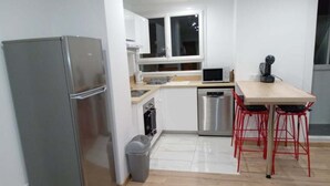 Eine kompakte Küche mit Kühlschrank, Mikrowelle und Geschirrspüler. An einem Holztisch stehen zwei rote Barhocker.