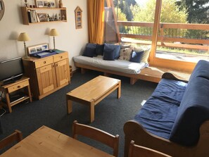 A cozy living room with a sofa, wooden furniture, a television, and a window with a view of trees.