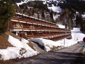 A wooden cabin with a sloped roof, situated on a snow-covered slope with a road leading up to it.