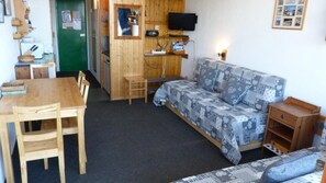 Een kleine, gezellige kamer met een houten tafel, een bank met een gestreept stoffering, een aan de muur bevestigde televisie, en een groene deur.
