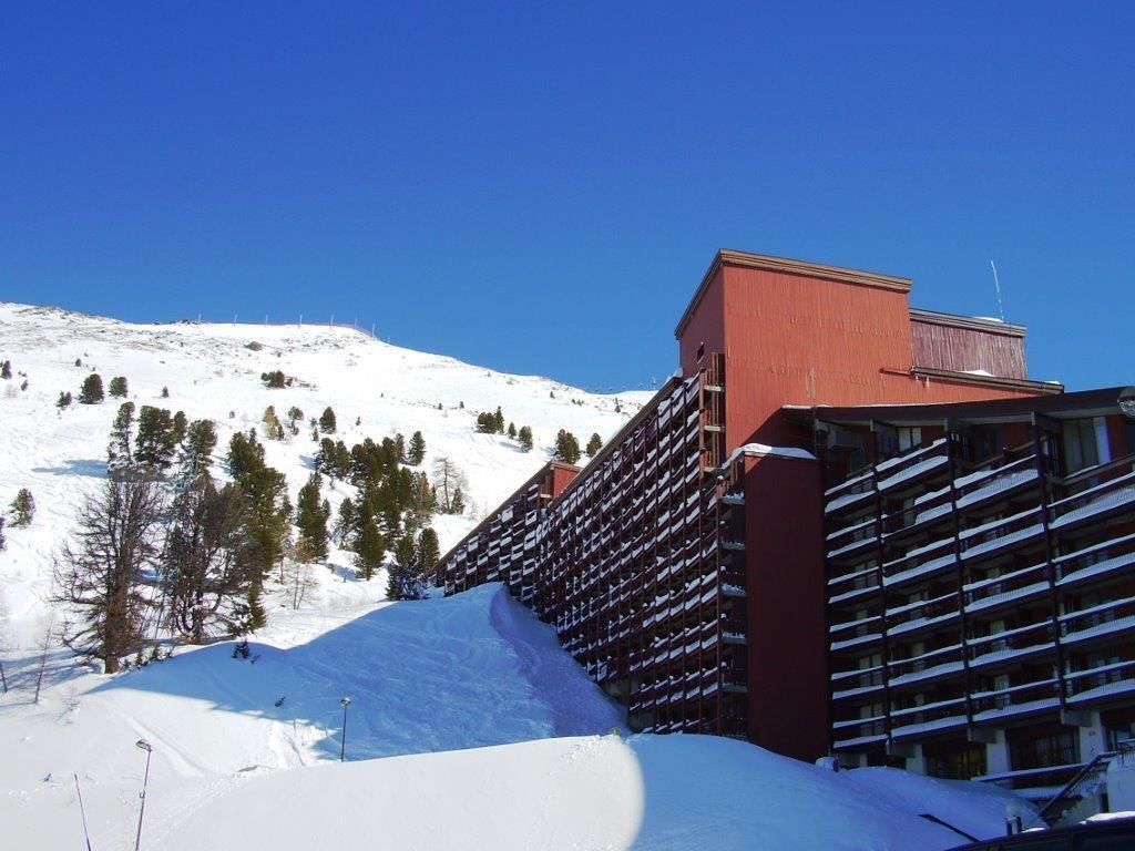 Ein mehrstöckiges Gebäude mit rotem Dach und Balkonen, umgeben von verschneiter Landschaft und Bäumen.