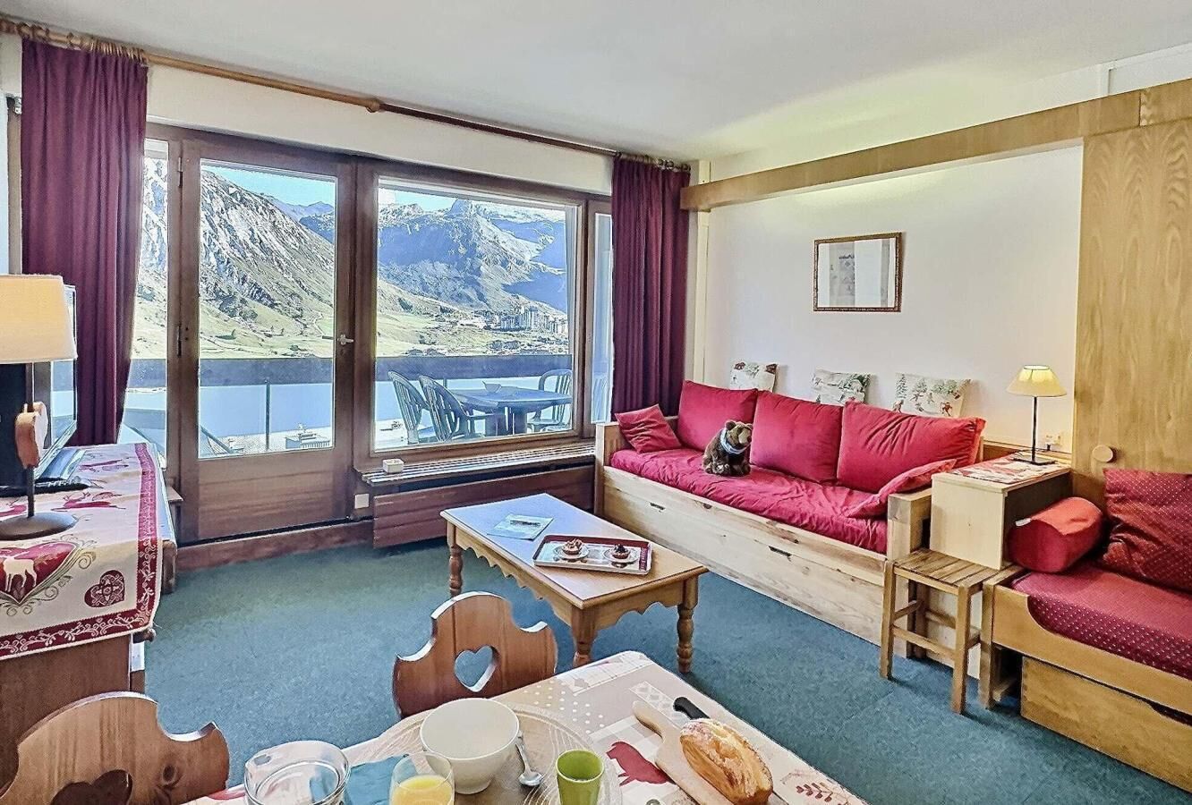 Quarto aconchegante com uma grande janela que oferece uma vista panorâmica das montanhas, um sofá de madeira com almofadas vermelhas e uma mesa de jantar com cadeiras.