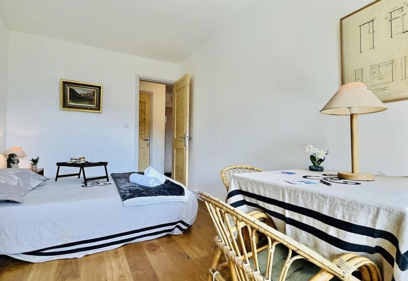 A room with two beds, a table, a chair, a lamp, and a framed picture on the wall.