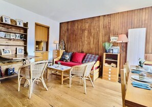 A cozy living room with a wooden wall, a red sofa, wicker chairs, and a dining table set for a meal.
