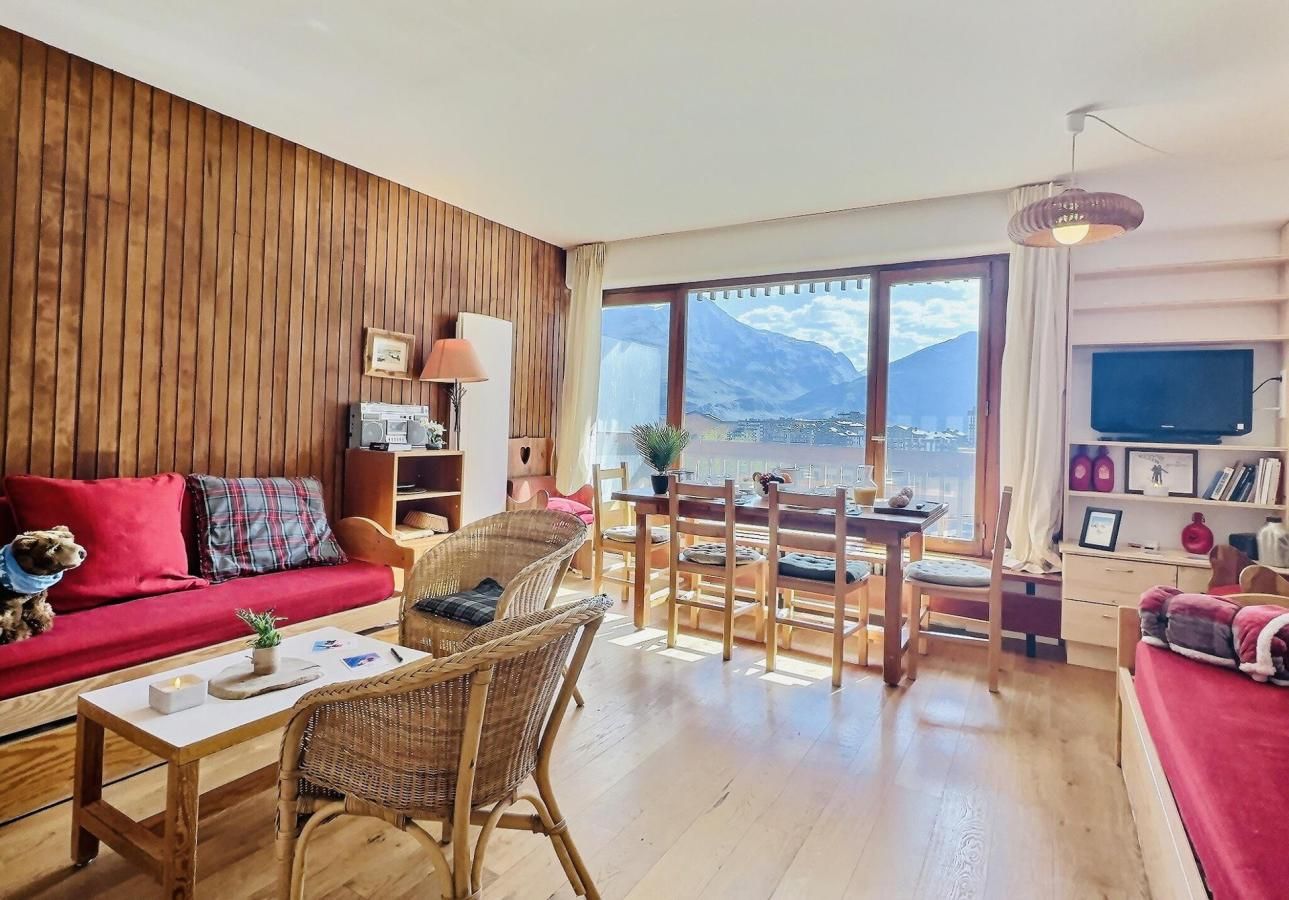 A cozy living room with a red sofa, wicker chairs, a wooden coffee table, and a large window offering a mountain view.