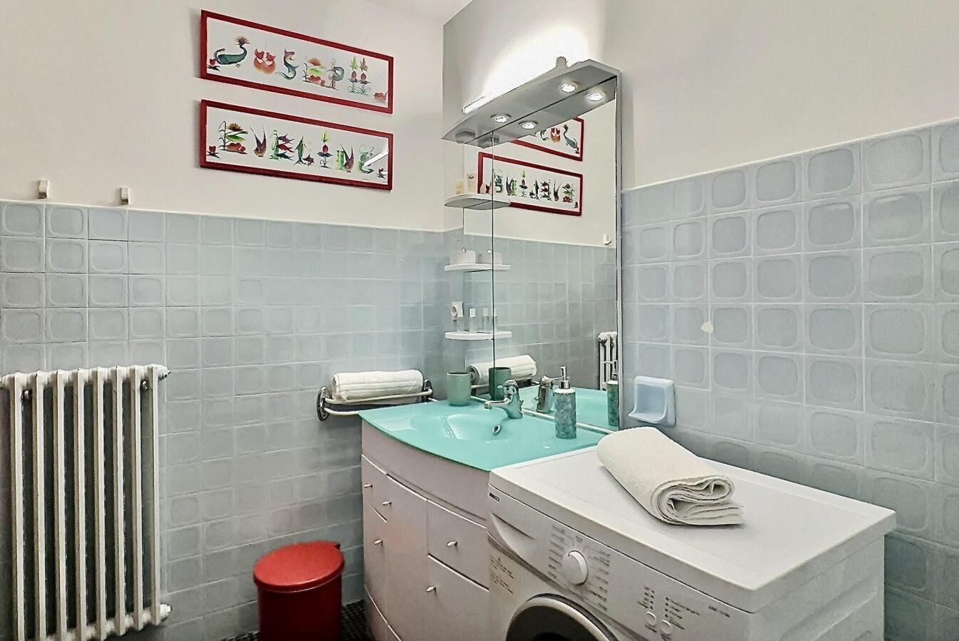 Ein Badezimmer mit Waschbecken, Spiegel und Heizkörper. Handtücher und ein roter Mülleimer sind vorhanden.