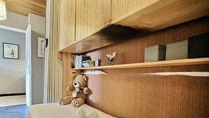 A neatly made bed with a teddy bear and rolled towels, wooden shelves above, and a framed picture on the wall.