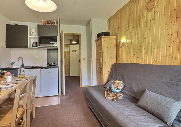 Un salon chaleureux avec un canapé gris, un meuble en bois et un ours en peluche. On aperçoit, au fond, une cuisine équipée d’un four à micro-ondes et d’une machine à café.