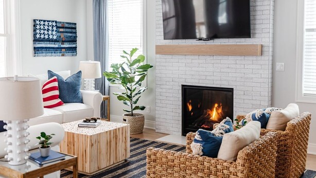 A living room with a white brick fireplace, a flat-screen TV mounted above it, a white sofa with blue and red striped pillows, a wicker armchair, and a coffee table with a plant.