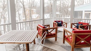 A covered outdoor seating area with wooden tables and chairs, large windows, and a view of a residential neighborhood.