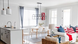 A modern kitchen with a central island, dining area with white chairs, a sofa with striped pillows, and a red 'LOVE' sign on the wall.