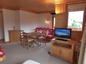 Um quarto acolhedor com uma mesa de jantar de madeira e cadeiras, um sofá vermelho, uma televisão sobre um suporte de madeira e uma janela com vista para edifícios.