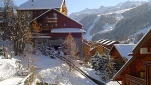 A wooden chalet-style building with a red roof, surrounded by snow-covered trees and mountains in the background. - 250m from ski slopes, 5th floor, view ski slopes, swimming pool, balcony, tv, ski locker, 32m² (Méribel - Les Allues)