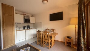 A compact living space with a kitchenette, a dining area with wooden chairs, a small table, a TV mounted on the wall, and a lamp on a side table. - 250m from ski slopes, 5th floor, view ski slopes, swimming pool, balcony, tv, ski locker, 32m² (Méribel - Les Allues)