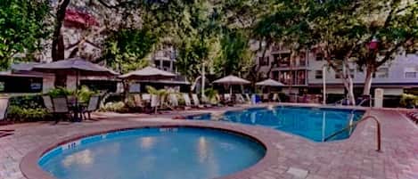 A swimming pool area with lounge chairs and umbrellas, surrounded by trees and buildings.