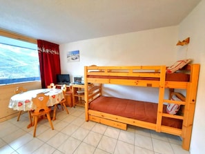A bunk bed with a desk and chair, a table with a heart-patterned tablecloth, and a window with a red curtain.