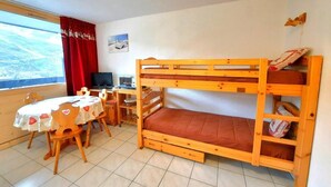 Uma cama beliche com escrivaninha e cadeira, uma mesa com toalha de coração e uma janela com cortina vermelha.