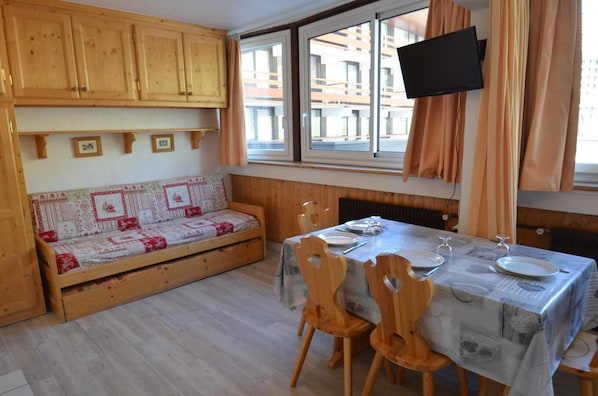 A cozy room with a wooden sofa, a dining table set for two, and a TV mounted on the wall.