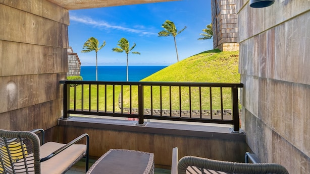 Un balcon avec des fauteuils en osier, une vue sur l’océan et une colline verdoyante parsemée de palmiers.