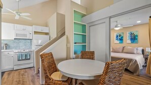 Une cuisine compacte avec des appareils électroménagers blancs, un coin repas avec des chaises en osier et une chambre avec un lit et des œuvres d’art.