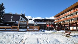 Une cour enneigée avec un banc en bois, une enseigne indiquant « LA TRATTORIA », et un bâtiment doté de balcons, avec un télésiège visible au loin.