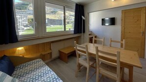 Une petite pièce comprenant un lit, une table en bois avec des chaises, un téléviseur et une fenêtre avec des rideaux.