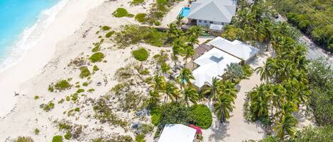 Vue aérienne d’une propriété située en bord de mer, avec une grande maison, une piscine et plusieurs palmiers.