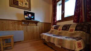 Una stanza accogliente con pavimento in legno, un divano con una coperta decorata, un televisore su una mensola di legno e una finestra con tende rosse.