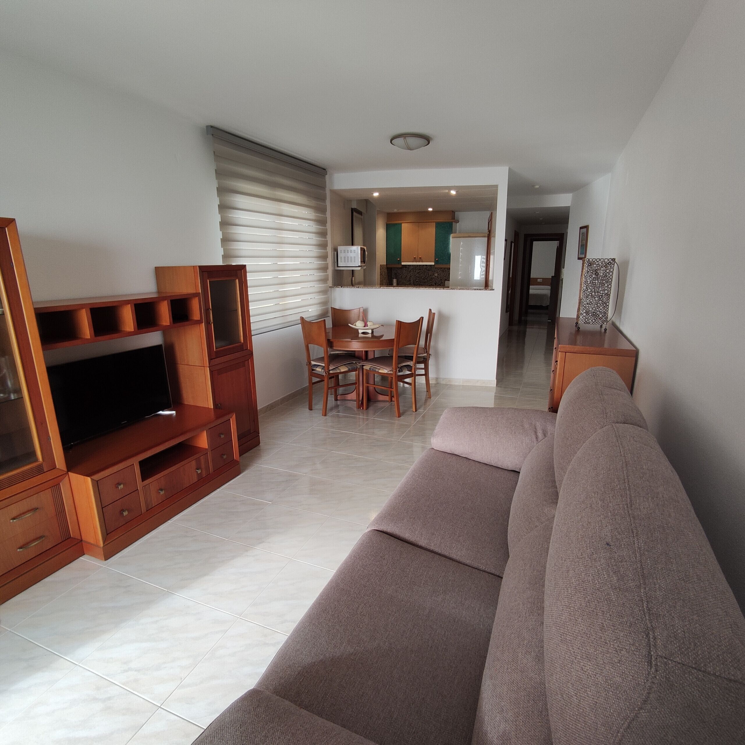 A living room with a sofa, TV, wooden entertainment unit, dining table with chairs, and a kitchen area with cabinets and a microwave.