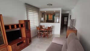 A living room with a sofa, TV, wooden entertainment unit, dining table with chairs, and a kitchen area with cabinets and a microwave. - Apartamento Da Vinci 5 Inmomart (Peñiscola)