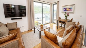 A spacious living room with a large TV, leather sofas, a wooden coffee table, and a dining area with a table and chairs. - 12 Loch View PF Kenmore (Aberfeldy)