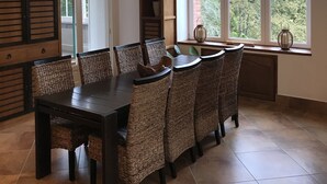 A dining area with a dark wooden table and wicker chairs, large windows with a view of trees, and a built-in bookshelf. - Château de La Tisnère on the outskirts of Pau (Gelos)