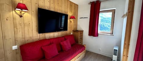 A room with a wooden wall, a red sofa, a flat-screen TV, a heater, and a window with red curtains.