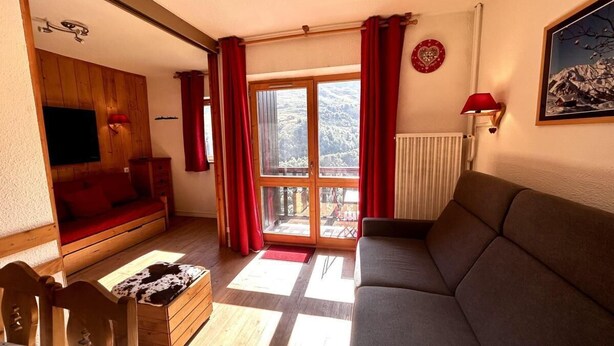 A cozy living room with a sofa, wooden furniture, a sliding glass door leading to a balcony, and a red curtain.