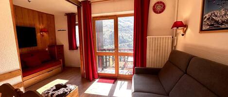 A cozy living room with a sofa, a wooden table, and a television. There is a sliding glass door leading to a balcony with a view of a snowy landscape.