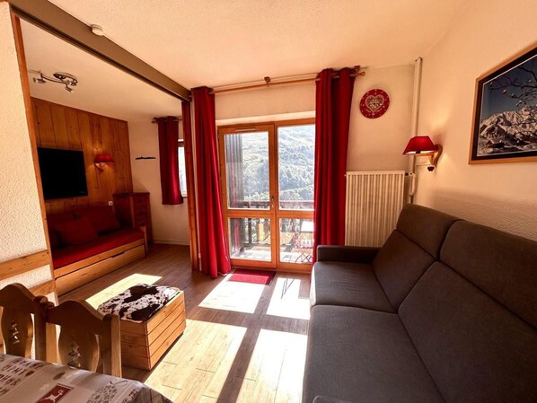 Una sala de estar acogedora con un sofá, una mesa de madera y un televisor. Hay una puerta corrediza de vidrio que da a un balcón con vistas a un paisaje nevado.