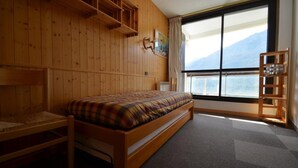 A wooden room with a bed, a small table, a shelf, and a large window offering a view of mountains. - 300m from resort center, 2nd floor, balcony, tv, 67m², Les Menuires (Les Menuires)