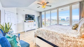 Camera da letto con un grande letto, una ventola a soffitto, una TV e vista sulla città attraverso ampie finestre.