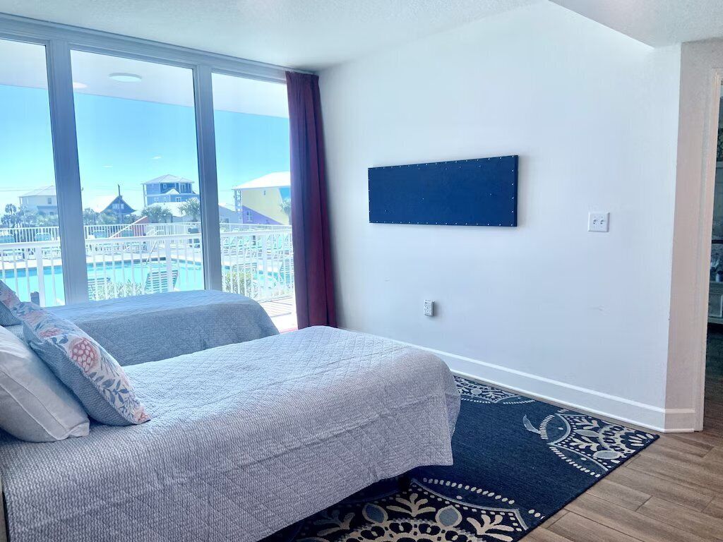 A bedroom with a bed, a large window, a view of buildings, and a mounted TV.