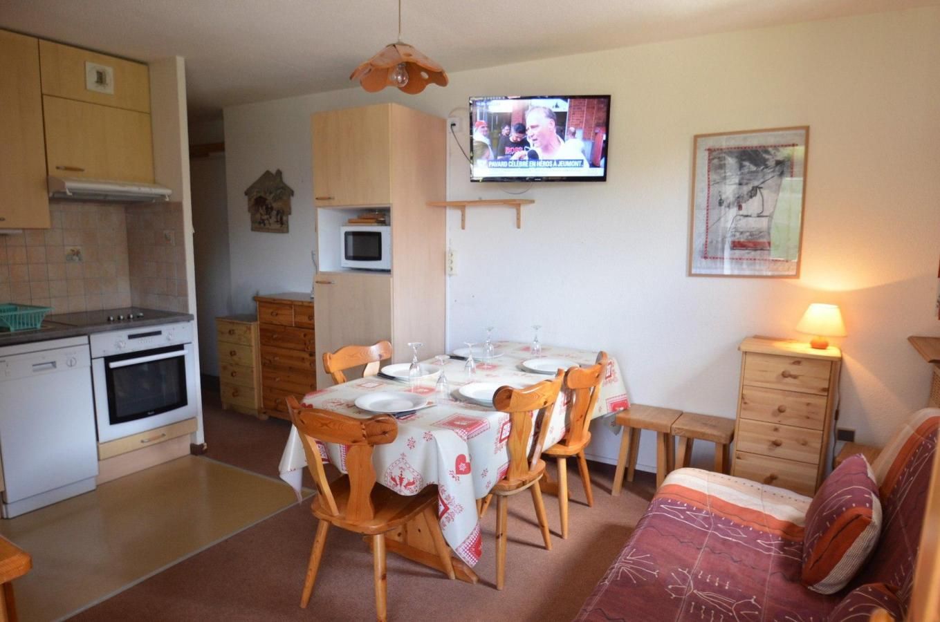 A compact kitchen with a microwave, oven, and wooden cabinets. A dining table with chairs is set for four, and a television is mounted on the wall.