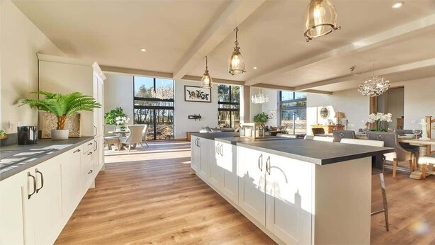 Een ruime keuken met een groot eiland, hangende lampen en uitzicht op het groen door grote ramen.