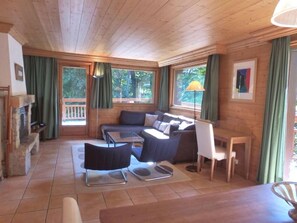 A cozy cabin interior with a stone fireplace, wooden ceiling, and large windows with curtains.