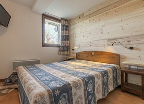 A bedroom with a bed, a nightstand, a wall-mounted TV, and a window with curtains.