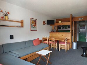 Un espacio habitable compacto con un sofá, una pequeña mesa de comedor con sillas, una zona de cocina con microondas y un televisor.